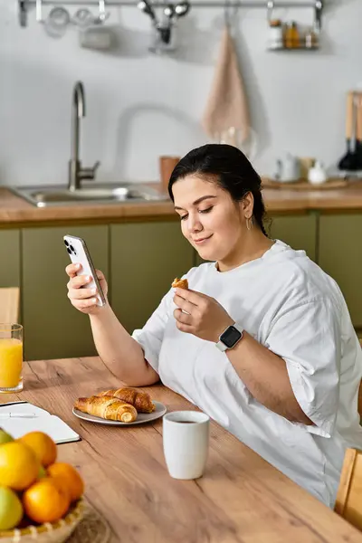 A lovely plus sized woman enjoys a croissant and juice while using her phone at breakfast. — Stock Photo