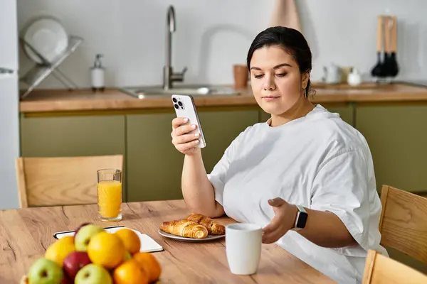A young plus sized woman savors her breakfast while checking her phone in a warm kitchen. — Stock Photo