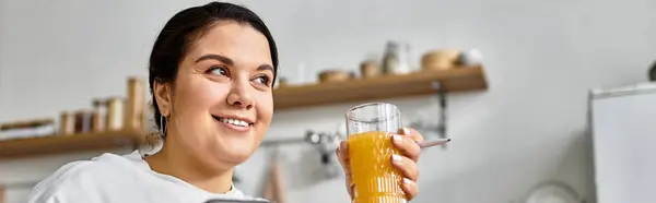 A young plus sized woman smiles happily with a colorful drink in a bright, sunlit kitchen. — Stock Photo