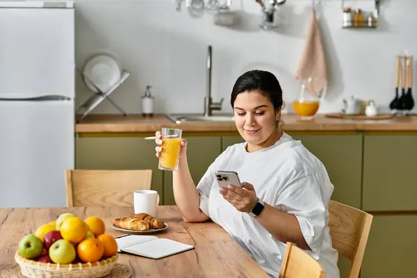 A young plus sized woman relaxes in her bright kitchen, sipping a drink while checking her phone — Stock Photo