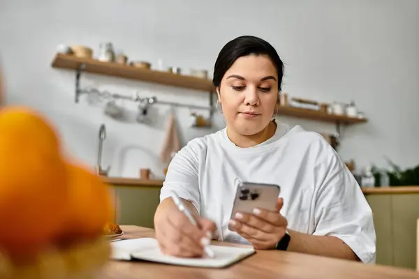 A young plus sized woman sits at a cafe table, jotting down notes while using her phone. — Stock Photo
