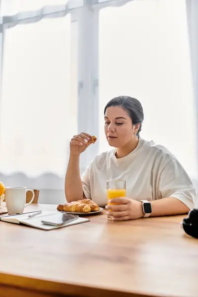 Savoring a morning meal, a young plus sized woman enjoys pastries and juice at a rustic table. — Stock Photo