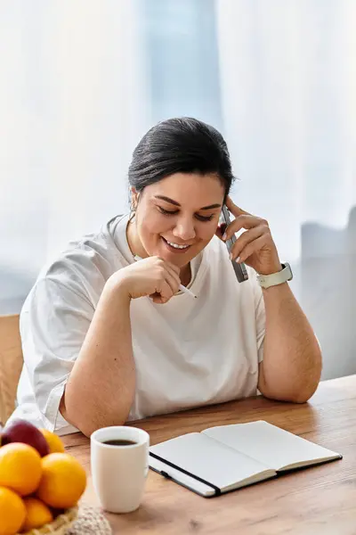 A smiling plus sized woman chats on the phone at a cozy wooden table with fruits. — Stock Photo