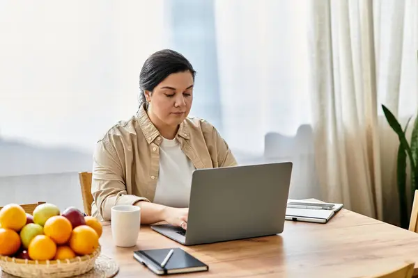 A beautiful plus sized woman works intently on her laptop at a wooden table with fruit. — Stock Photo