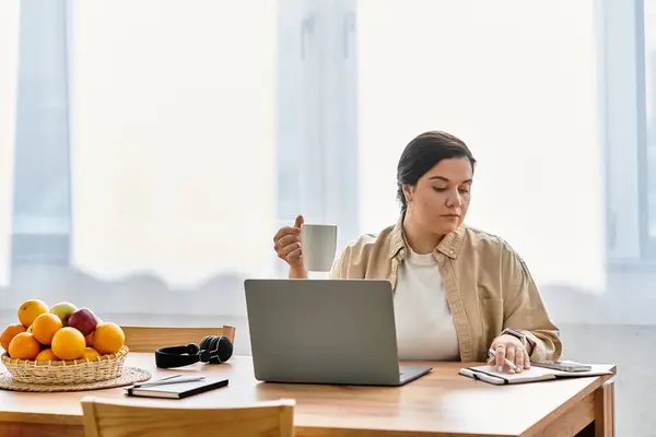 Plus sized woman sits at a wooden table sipping tea and focusing on her laptop in a sunlit space. — Stock Photo