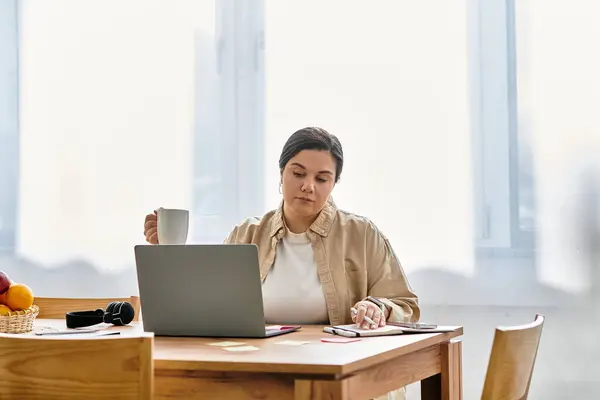 Bright sunlight fills the room as a young plus sized woman focuses on her work with a warm drink. — Stock Photo