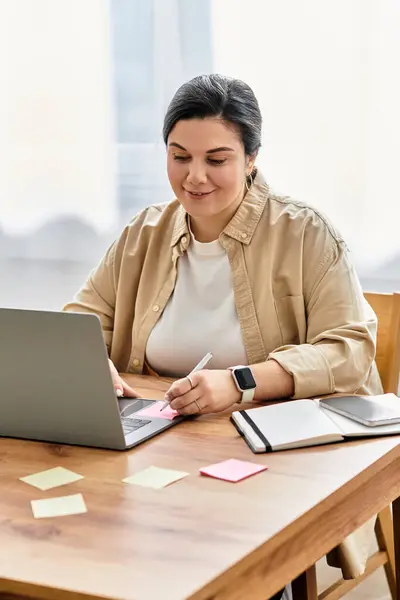 Une jeune femme de taille plus s'assoit à son bureau, en toute confiance en utilisant son ordinateur portable tout en prenant des notes. — Photo de stock