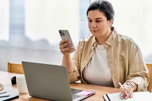 A young plus sized woman sits at a wooden table, focused on her phone while working on her laptop. — Stock Photo
