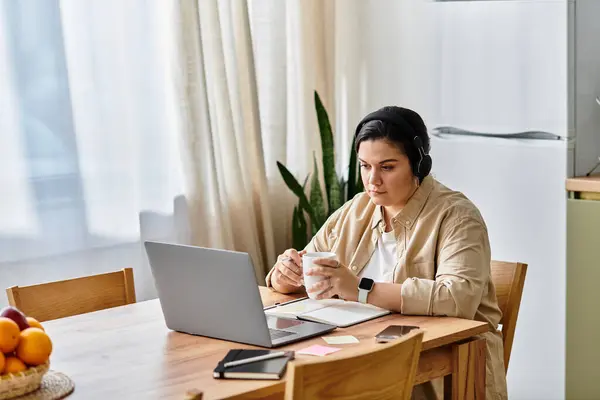Beautiful young woman with headphones enjoys a warm drink while working on a laptop. — Stock Photo