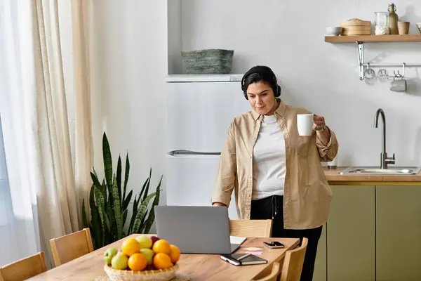 A lovely plus sized woman sips a warm drink while working at a cheerful kitchen table. — Stock Photo