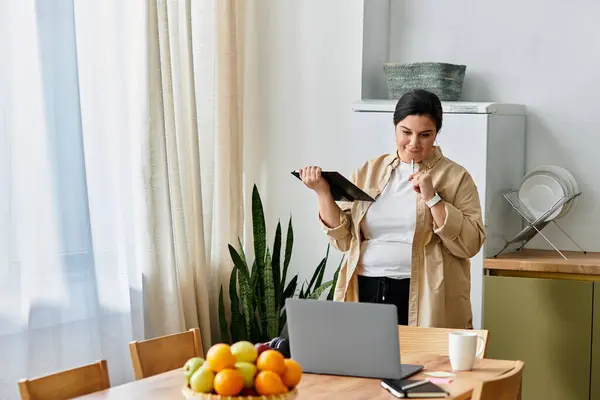 Uma jovem mulher de tamanho grande sorri enquanto trabalhava em um laptop em sua cozinha acolhedora cheia de frutas. — Fotografia de Stock