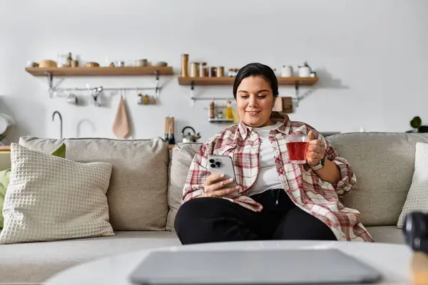 A young plus sized woman relaxes on a sofa, sipping tea while checking her smartphone. — Stock Photo