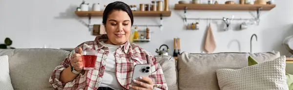 A plus sized woman relaxes on the couch, sipping tea and using her phone. — Stock Photo