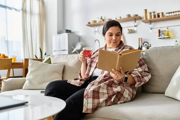A young plus sized woman relaxes on a sofa, sipping tea while immersed in an engaging book. — Stock Photo