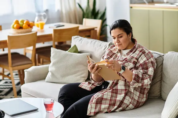 A young plus sized woman sits comfortably on a couch reading a book, immersed in her thoughts. — Stock Photo