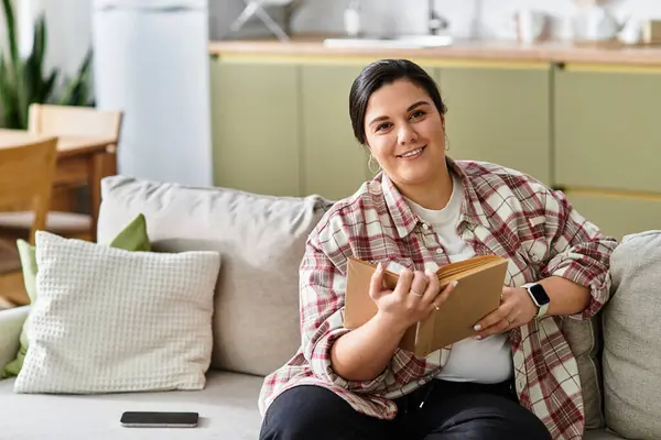 Young plus sized woman smiles warmly while reading a book in her stylish, sunny living room. — Stock Photo