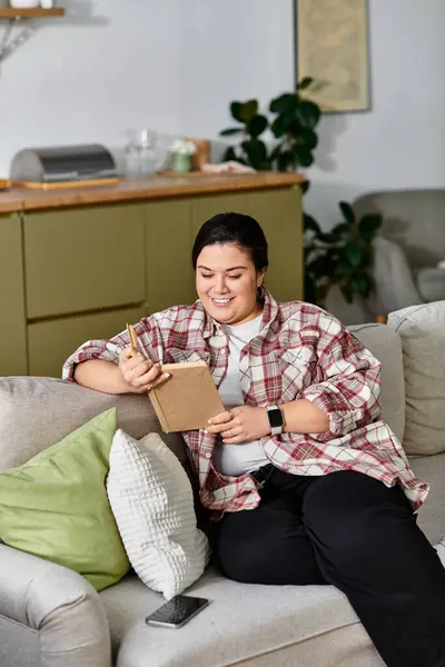 Uma mulher sorrindo plus size escreve em seu caderno em um ambiente acolhedor em casa. — Fotografia de Stock