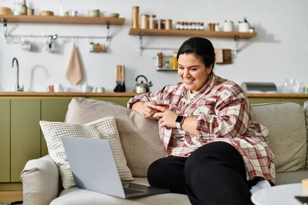 A vibrant and cozy setting features a young plus sized woman sipping tea while using her laptop. — Stock Photo