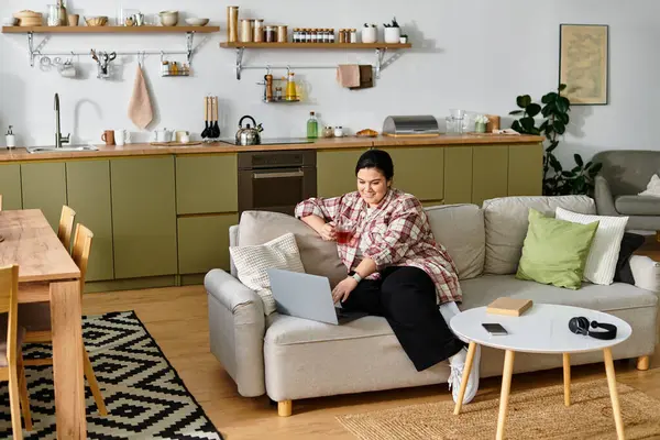 A young plus sized woman relaxes on a couch, sipping a drink and using her laptop. — Stock Photo