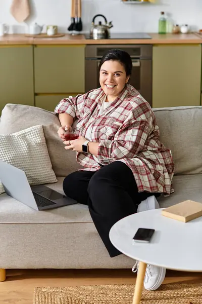 Uma mulher jovem, bonita e de tamanho grande senta-se confortavelmente em um sofá, bebendo uma bebida e sorrindo. — Fotografia de Stock