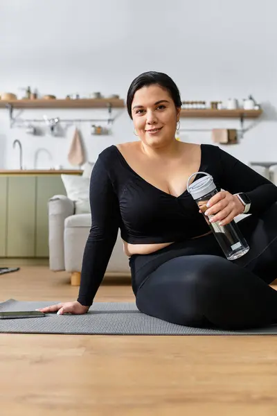 Una joven mujer de tamaño grande se relaja con una botella de agua después de completar su rutina de ejercicios en casa. - foto de stock