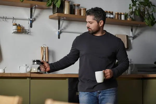 A handsome man stands in a modern kitchen, pouring coffee into a mug with a calm expression. — Stock Photo