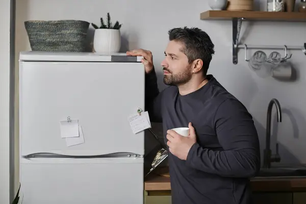 A man thoughtfully holds a cup by the fridge in a modern kitchen. — Stock Photo