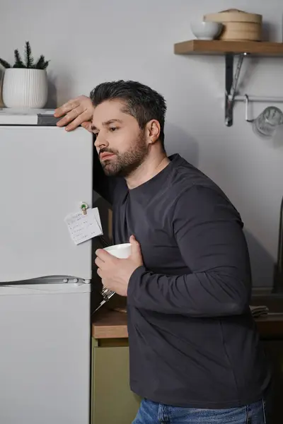 Um homem se inclina contra a geladeira, refletindo pensativamente enquanto segura uma xícara em uma cozinha elegante. — Fotografia de Stock