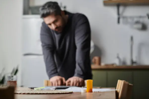 Um homem bonito inclina-se para a frente pensativo em uma mesa em uma cozinha quente, foco em pílulas. — Fotografia de Stock