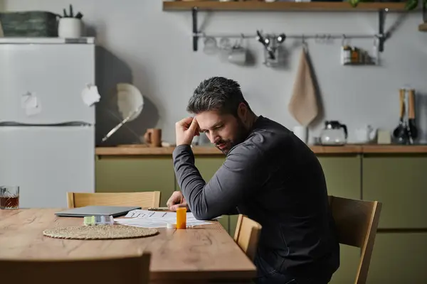 Un hombre guapo se ve contemplativo mientras examina los papeles en una cocina cálida y acogedora. - foto de stock