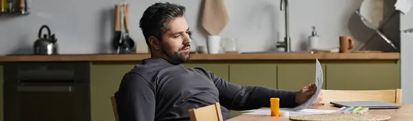 Um homem encantador senta-se confortavelmente em uma mesa de cozinha, examinando papéis com foco e facilidade. — Fotografia de Stock