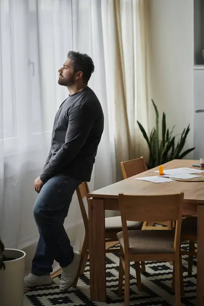 Un bel homme se tient près de la fenêtre, perdu dans la pensée, tandis que la lumière du soleil remplit l'espace confortable. — Photo de stock