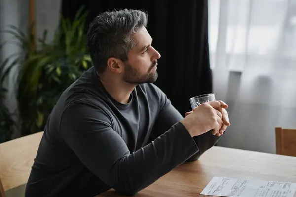 Un bell'uomo si siede pensieroso a un tavolo di legno, tenendo un drink mentre guarda fuori da una finestra. — Foto stock