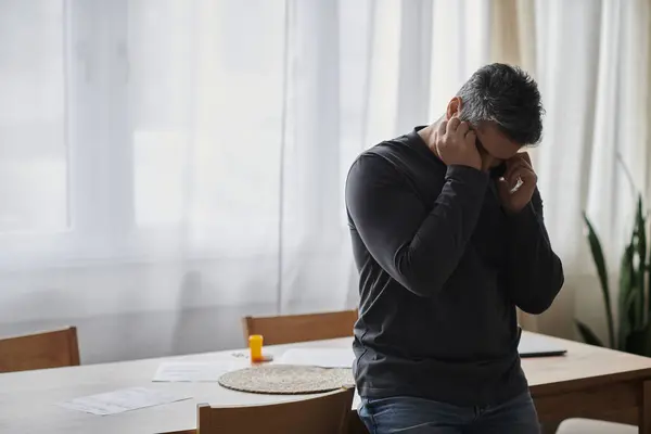 Un hombre guapo parece considerado mientras habla por teléfono en una habitación luminosa y acogedora.. - foto de stock