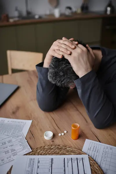 Un bell'uomo siede da solo a un tavolo rustico, sopraffatto da medicine e carte sparse. — Foto stock