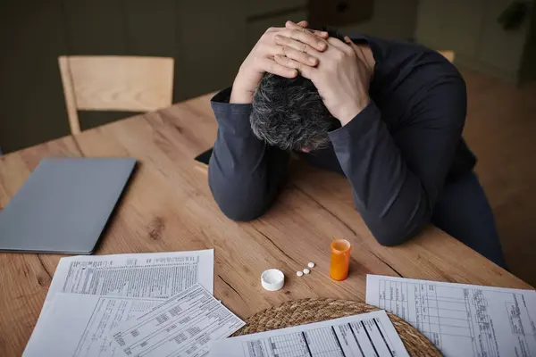 A handsome man at a wooden table looks stressed, grasping his head while reviewing papers. — Stock Photo
