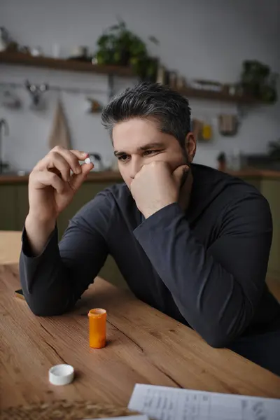 A handsome man at a wooden table thoughtfully examines a pill in a cozy kitchen. — Stock Photo