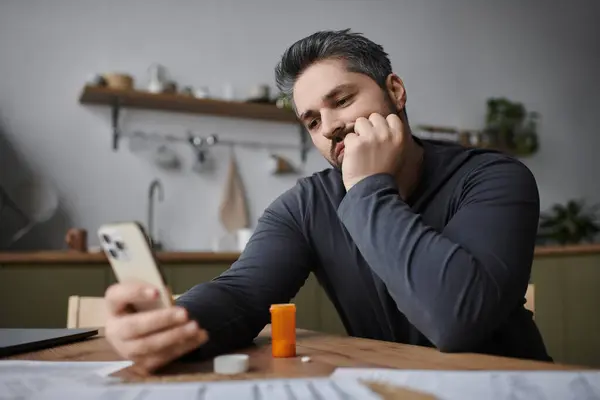 Un hombre guapo apoya su barbilla en su mano, inmerso en su teléfono inteligente, rodeado de comodidades para el hogar. - foto de stock