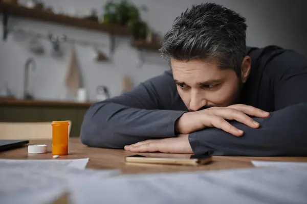 A thoughtful man leans on his arms at a table filled with papers and a bottle of medication. — Stock Photo