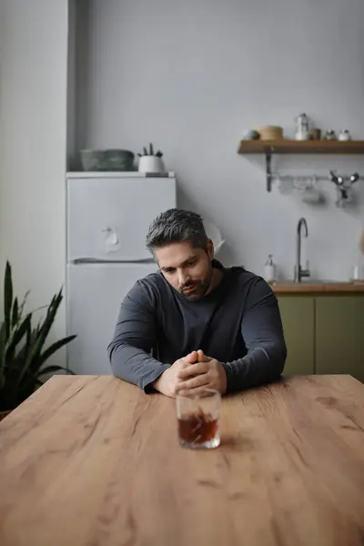 A handsome man sits at a wooden table, staring at a drink in a modern kitchen. — Stock Photo