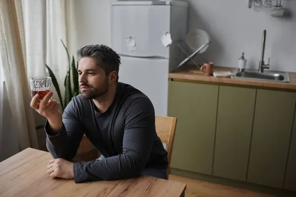 A handsome man sits at a wooden table, thoughtfully examining a glass of whiskey in a cozy kitchen. — Stock Photo