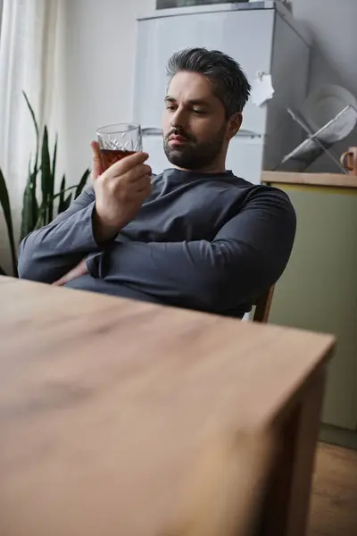 A handsome man sits at a wooden table, contemplating life as he examines his drink in a serene home. — Stock Photo