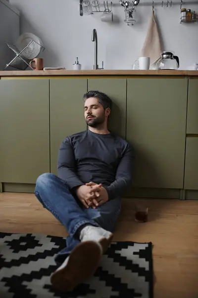 A handsome man sits peacefully on the kitchen floor, lost in thought while sipping a drink. — Stock Photo
