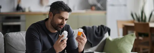 Un bel homme s'assoit confortablement sur le canapé, analysant deux flacons de médicaments avec attention. — Stock Photo