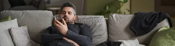 A handsome man lounges on a comfortable couch, absorbed in his phone and savoring a drink. — Stock Photo