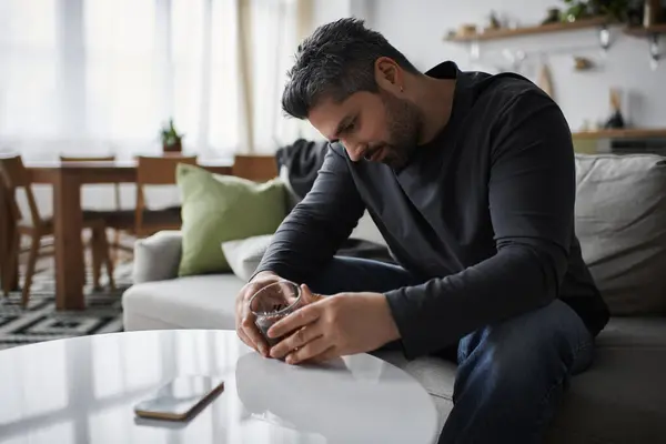A handsome man sits on a couch with a drink, surrounded by soft decor and natural light. — Stock Photo