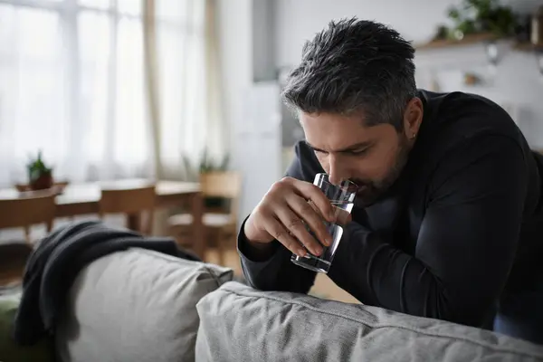A handsome man leans forward, lost in thought while holding a glass of water in his living room. — Stock Photo