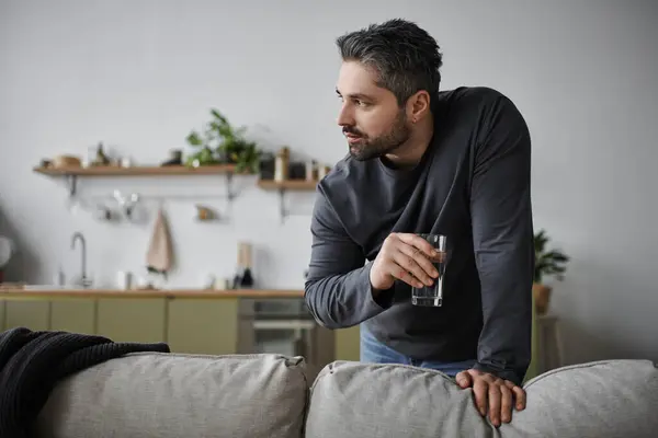 El hombre reflexivo se apoya en un sofá mientras sostiene un vaso de agua en una cocina elegante. - foto de stock