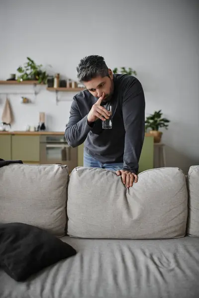 A handsome man is leaning on a sofa, sipping a drink, and contemplating life in a modern home. — Stock Photo