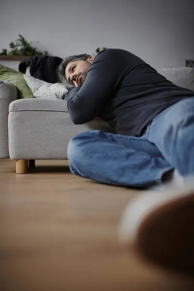 A handsome man lies on the floor, wrapped in thoughts, near a comfortable sofa and soft pillows. — Stock Photo